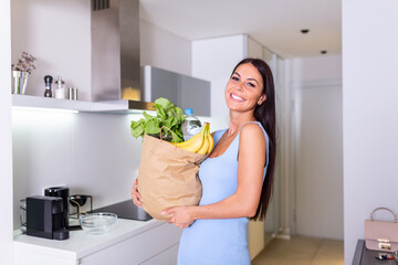 Young woman holding grocery shopping bag with vegetables Standing in the kitchen. Happy Woman Holding Grocery Shopping Bag With Vegetables In Kitchen At Home