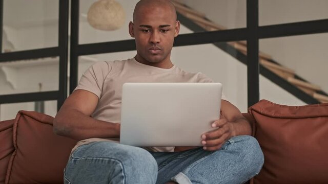 A focused concentrated african american man is using his laptop sitting on the couch in living room at home