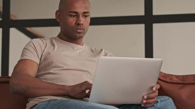 A focused concentrated african american man is using his laptop sitting on the sofa in living room at home