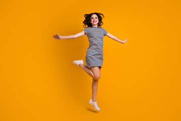 Full body photo of funky curly lady jump high spread hands sides weekend rejoicing sunny day wear white casual striped short dress footwear isolated yellow color background