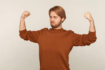 Fototapeta premium I am strong! Portrait of confident proud bearded man in sweatshirt demonstrating power in his hands, showing biceps, feeling energy to win success. indoor studio shot isolated on gray background