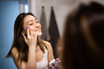 Portrait of young asian woman applying moisturizer cream on her pretty face and look mirror.