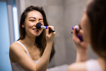 Beautiful young asian woman applies makeup in front of a bathroom mirror