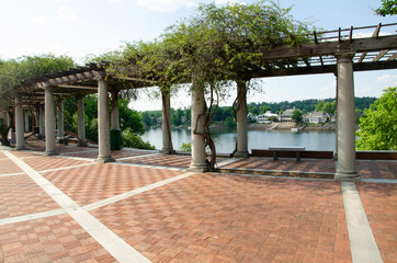 A section of a river walk with a shaded rest area under a pergola, with white columns. There is a view of the river.