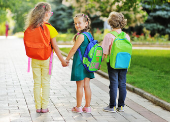 three school children-two girls and a curly-haired boy with school colored bags smiling hold hands and go to school. Back to school concept, September 1, knowledge day, education.