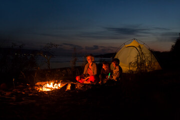 Family mom and two kids sitting together around campfire, enjoying fresh air near tent on the seashore. Tourism, camping concept. Staycations, hyper-local travel,  family outing, getaway.