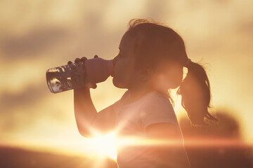 Caucasian child girl re-hydrating drinking some water against a sun flare. Outdoor scene with...
