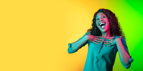 Smiling, looks shy and happy. African-american young woman's portrait on gradient background in neon. Beautiful female model. Concept of cinema, emotions, facial expression, sales, ad. Flyer.