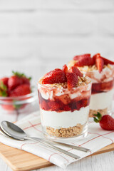 Homemade layered dessert with fresh strawberries, cream cheese or yogurt, granola and strawberry jam in glasses on white wood background. Healthy organic breakfast or snack concept. Selective focus.