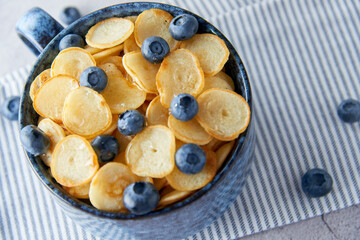 Blue Bowl of mini pancake cereal with blueberries, maple syrup and milk on concrete background. Tiny pancakes, new food trend concept. Cute healthy breakfast or snack. Recipe, menu.