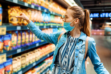 young woman in a medical protective mask on her face buys food at a supermarket. a young lady...