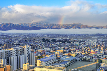 A bird's-eye view of a hotel in Yamagata City that overlooks the surrounding town, with densely-covered mountains and rainbow appearing as a beautiful nature after the rain.