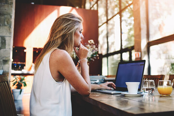 Portrait of a young beautiful woman working on laptop computer while sitting in modern cafe bar interior, blonde female freelancer using net-book for remote job during morning breakfast in coffee shop