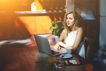 Pretty female freelancer using laptop computer for creating new project while sitting in co-working cafe, beautiful Sweden woman chatting with friends in social network via net-book during rest in bar