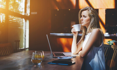 Young charming woman dreaming about something while enjoying good tea after work on laptop computer, thoughtful hipster girl drinking coffee while waiting answer on her e-mail on portable net-book