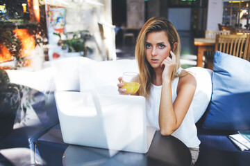 Lovely woman holding glass of orange juice while sitting front open laptop computer in comfortable coffee shop interior, young Sweden female posing during work on portable net-book in cozy cafe-bar