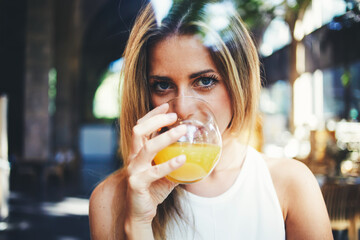 View though the window of a young gorgeous woman enjoying the glass of fresh orange juice in modern coffee shop interior, attractive female drinking natural vitamin beverage and looking at you