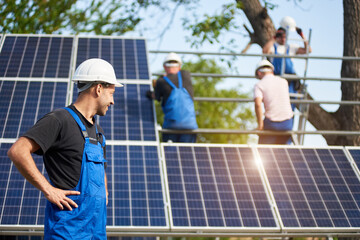 Profile of successful engineer technician standing in front of unfinished high exterior solar panel photo voltaic system blue shiny surface, looking at team of workers on high platform.