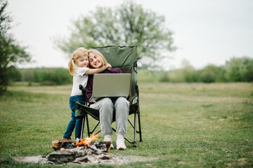 Plakat Mother work on Internet with kid outdoors. Quarantine, closed nursery school during coronavirus outbreak. Communication with family online on laptop near fire in nature. Homeschooling, freelance job.