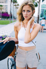 Half length portrait of young charming blonde hair woman standing with her classic bicycle in the street while looking at you,wonderful female cyclist dressed in trendy clothes enjoying summer weekend