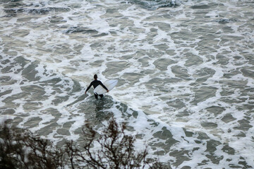 an unidentifiable lone male surfer carrying his surfboard over the rocks of the beach into the churning crashing waves on a rainy surf day, Bell's beach, great ocean road, Victoria, Australia