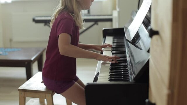 A beautiful blond little girl in a burgundy T-shirt and shorts sits behind the piano and diligently play a note. View from the side. Close-up