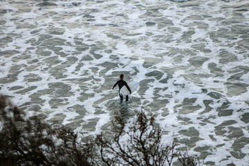 an unidentifiable lone male surfer carrying his surfboard over the rocks of the beach into the churning crashing waves on a rainy surf day, Bell's beach, great ocean road, Victoria, Australia