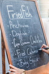 Mano escribiendo con un rotulador sobre una pizarra platos de tipica comida mediterránea escrita...