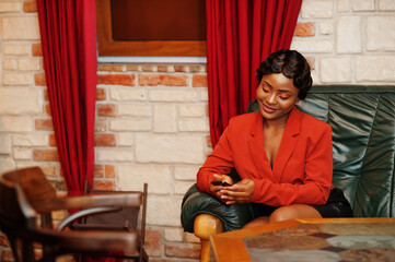 Portrait of african american woman, retro hairstyle,sitting on couch with mobile phone,  wear orange jacket and leather skirt posing at restaurant.