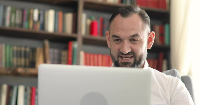 Portrait of man is talking video call sitting in office on bookshelves background. Remote work of teacher, professor or entrepreneur online at the computer, laptop in office. Remote work concept.