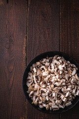 Tasty chopped champignon mushrooms in frying pan, uncooked food sliced, dark brown wooden background, top view copy space