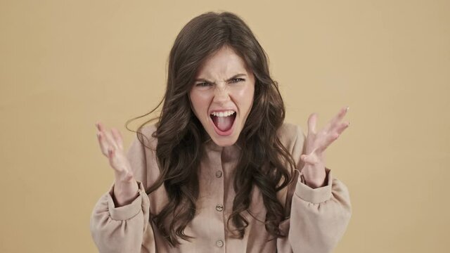An angry woman is screaming and clutching her head isolated over a beige background