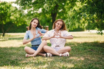 Fototapeta premium Two beautiful girls have fun in the park on a sunny summer day. The sisters are sitting on a green lawn, laughing.