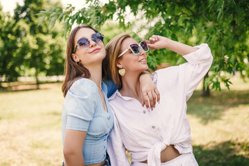 Two beautiful girls spring in the park on a sunny summer day. Sisters walk in fashionable outfits in sunglasses.