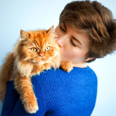 Portrait of beautiful short haired 30 years old woman in blue dress kissing her ginger red furry persian cat. Domestic animal, pet and host together. People love kitten. Cat day