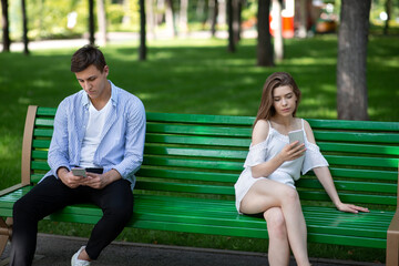 Gadget addiction and relationship problems. Young couple with smartphones sitting on bench at park,...