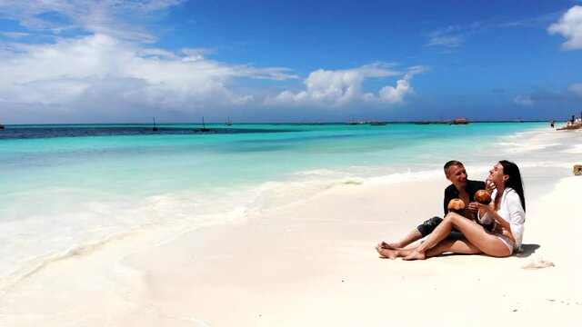Unforgettable honeymoon of young couple in love, pretty wife in white bikini sitting on white sand and tasting exotic beverage. Summer relax at the maldives resort, couple enjoying summer vacations