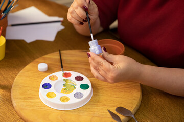 Fototapeta na wymiar Close up female artist's hands mixes colors on a paint palette on wooden table in art home studio workspace. Handmade art craft, DIY, workshop, decoration painting utensils, creative hobby concept
