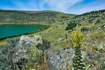 Emerald Lake Nar in the crater of an extinct volcano. Summer sunny day.On the slopes of the mountains around the round lake are picturesque boulders. Wildflowers and trees are growing. Bright blue sky