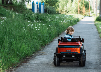 curly-haired boy in a striped T-shirt rides a red big toy car driving on an asphalt path. day off, outdoor recreation