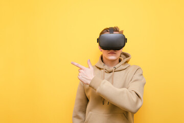 A young man wears a VR helmet and points his finger away at a blank space, isolated on a yellow...
