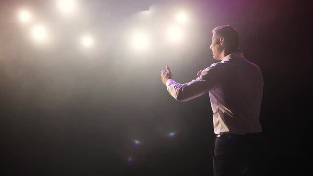 Man stand up comedian speaking jokes in micropphone headset standing on stage, back view. Guy showman entertains the audience under the spotlights. Concert, show, performance concept.