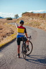 Fototapeta premium Young cyclist woman standing on her bike in the middle of the road on her back while pointing at a point of the landscape.