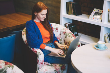 Smiling copywriter with red hair keyboarding information on laptop computer working remote in...