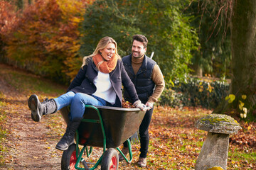 Man Pushing Woman In Wheelbarrow As Couple Rake Autumn Leaves From Garden Together
