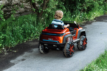 curly-haired boy in a striped T-shirt rides a red big toy car driving on an asphalt path. day off, outdoor recreation