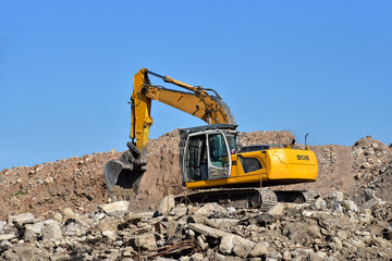 Yellow excavator at landfill for disposal of construction waste. Backhoe dig gravel at mining quarry on blue sky background. Recycling concrete and asphalt from demolition.