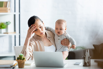 selective focus of upset mother looking at laptop and holding infant son