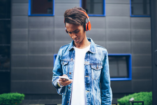 Stylish hindu hipster listening music in modern headphones choosing song in own playlist on telephone standing on promotional background.Serious male reading text message with bad news on cellular
