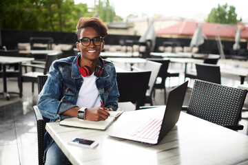 Half length portrait of happy hindu hipster student with headphones on neck doing homework in notepad during studying at modern laptop device sitting outdoors in cafe and using free wifi connection